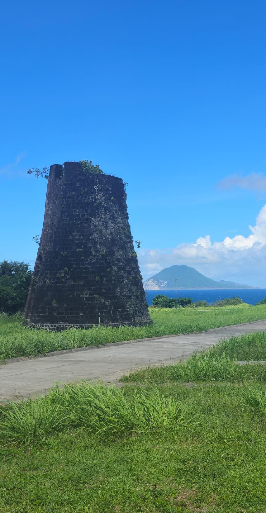 Weathered tapered stone tower overgrown with plants beside a paved path, with the sea beyond